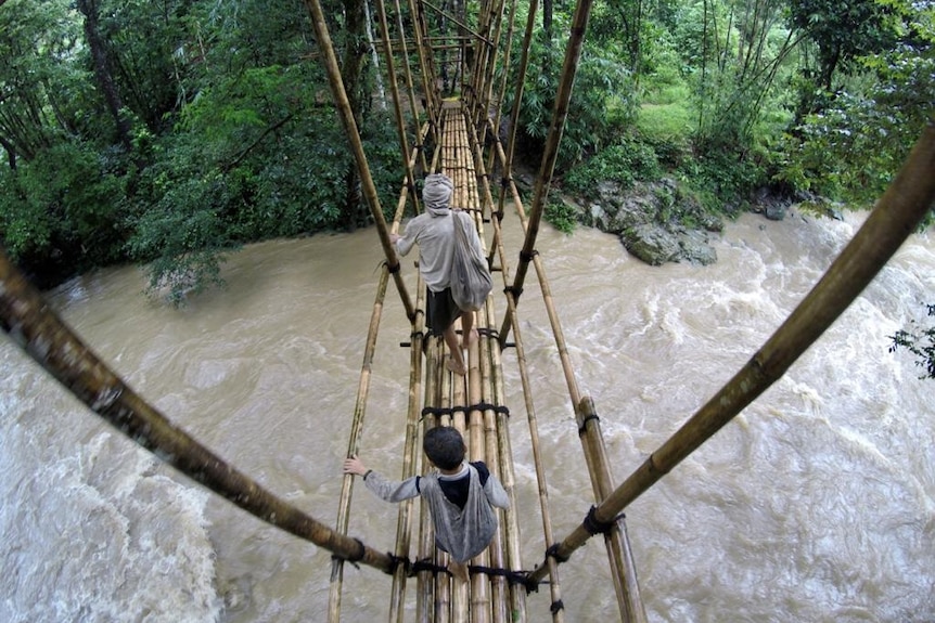 Two men crossing a bridge made from a bamboo with a river flowing underneath