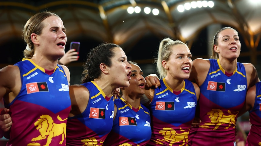 Brisbane Lions players stand arm-in-arm next to each other singing