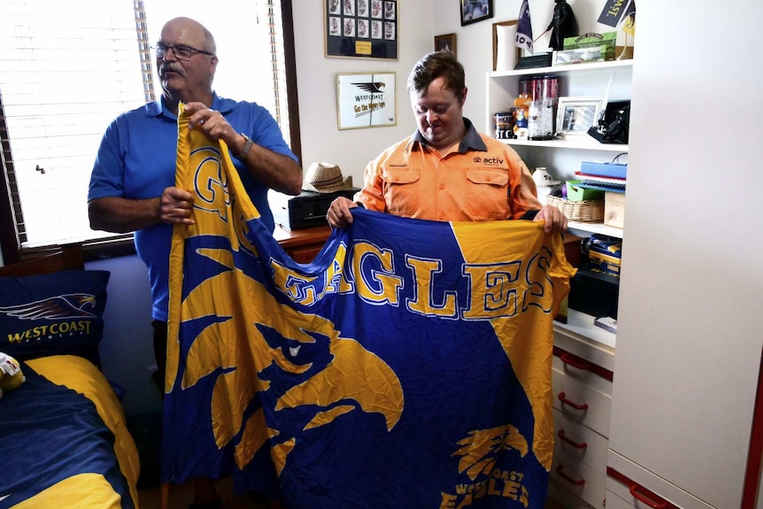 Two men stand together in a bedroom holding a AFL team West Coast Eagles flag