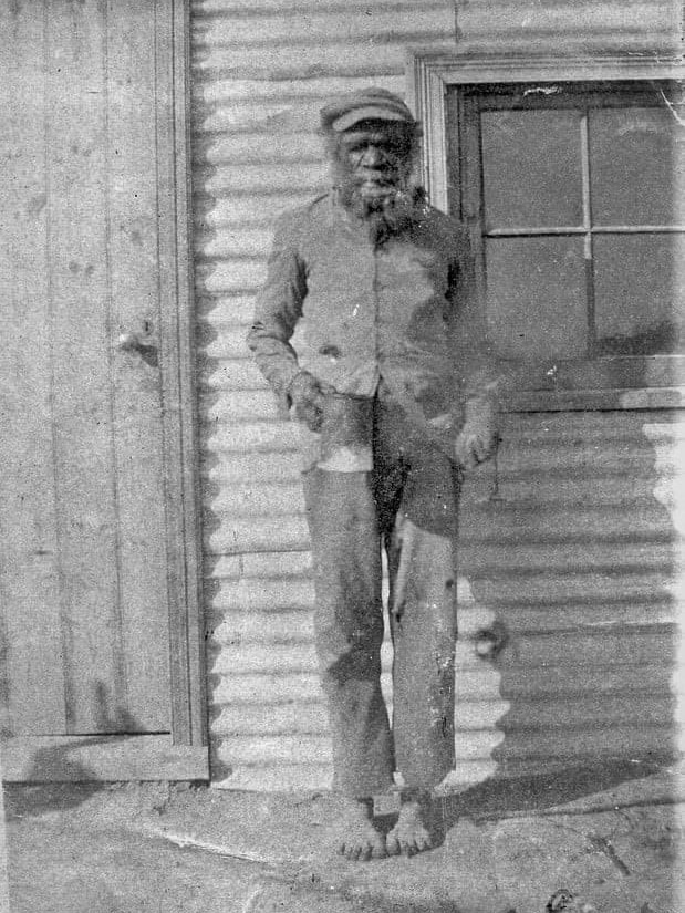 He wears worn clothes, a cap on his head, bare feet and holds a mug. The photo is old and black and white