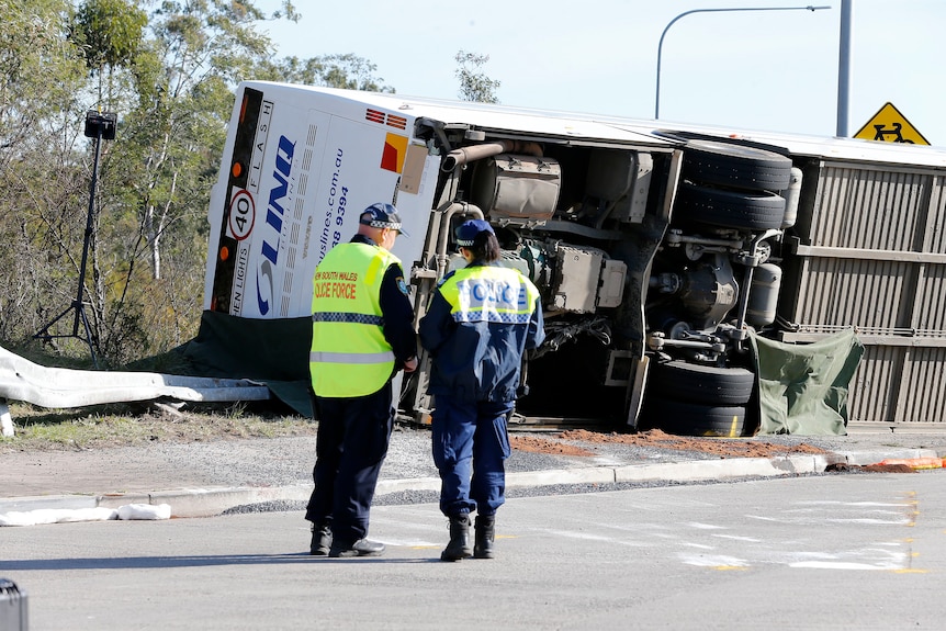 Two police officers in uniform stand and look at the bottom of an overturned bus