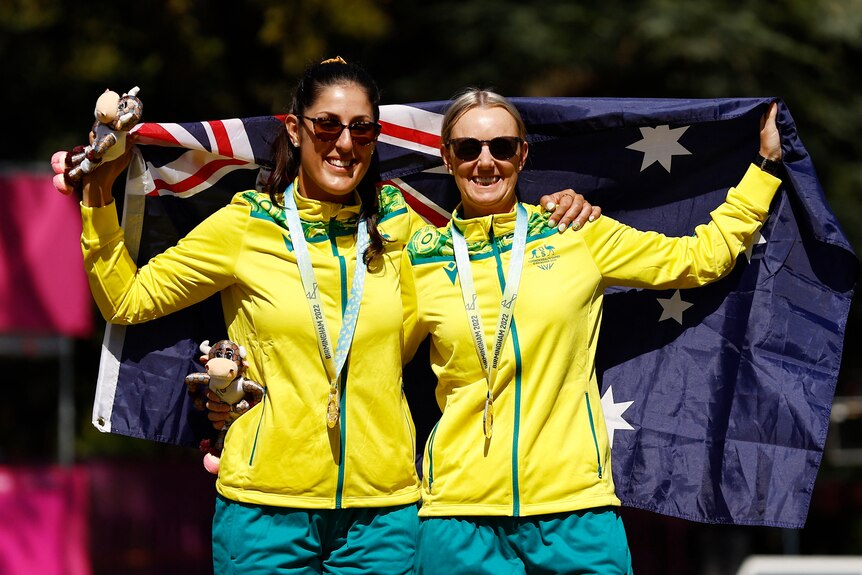 two young australian lawn bowls players smile with their arms around each other draped in australian flag wearing gold medals