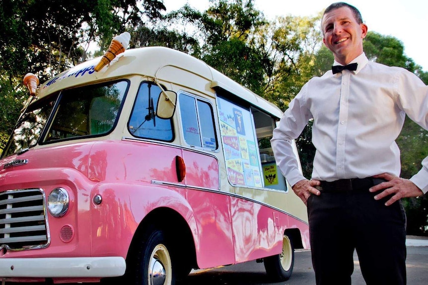 A man in a white shirt wearing a bow tie stands with his hands on his hips next to a pink ice cream van.