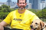 A smiling, dark-haired man with a salt-and-pepper beard, wearing a bright T-shirt and sitting with his dog.