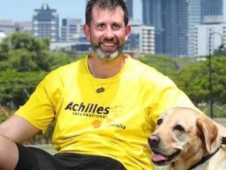 A smiling, dark-haired man with a salt-and-pepper beard, wearing a bright T-shirt and sitting with his dog.