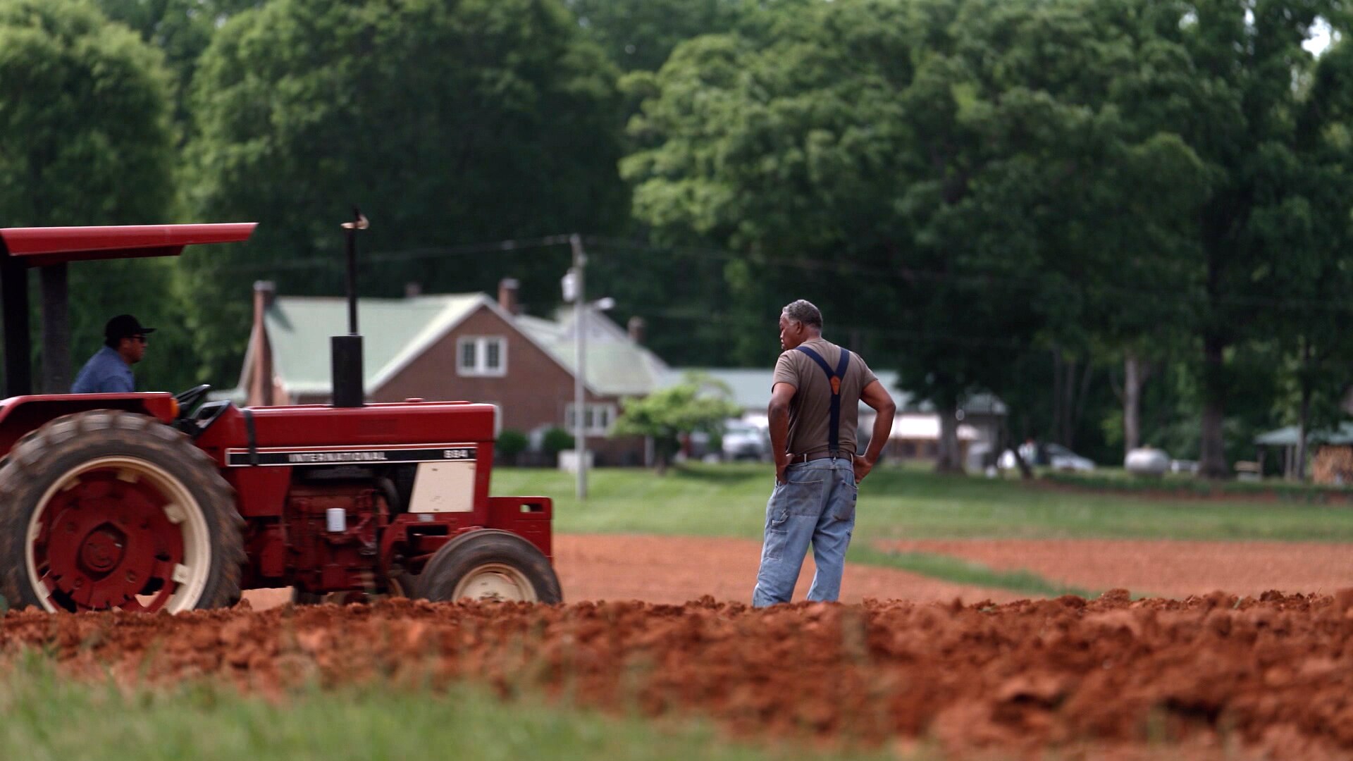 A man stands, his hands on his hips, in the middle of a field of a farm. A tractor is passing him.