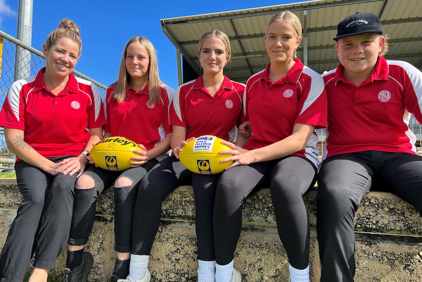 A mum and her four teenage children sit on a bench, with two yellow AFL footballs.