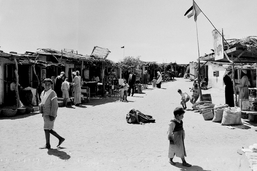 Black and white photo of children and adults walking through refugee camp. Small girl naps in the middle of the street
