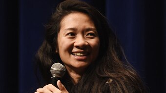 Chinese-American woman late 30s with long hair, smiling and holding mic, speaking.