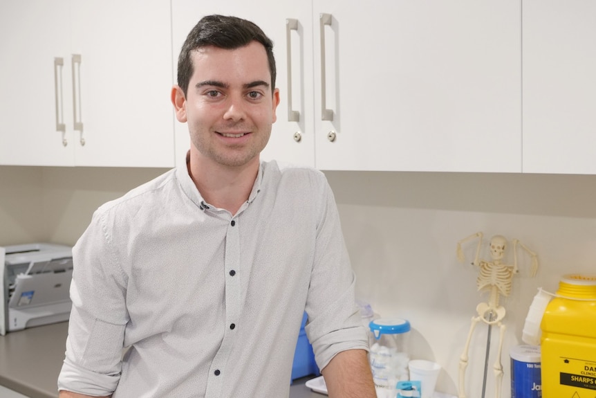 A man in a button up shirt leaning on a bench with medical utensils in the background.