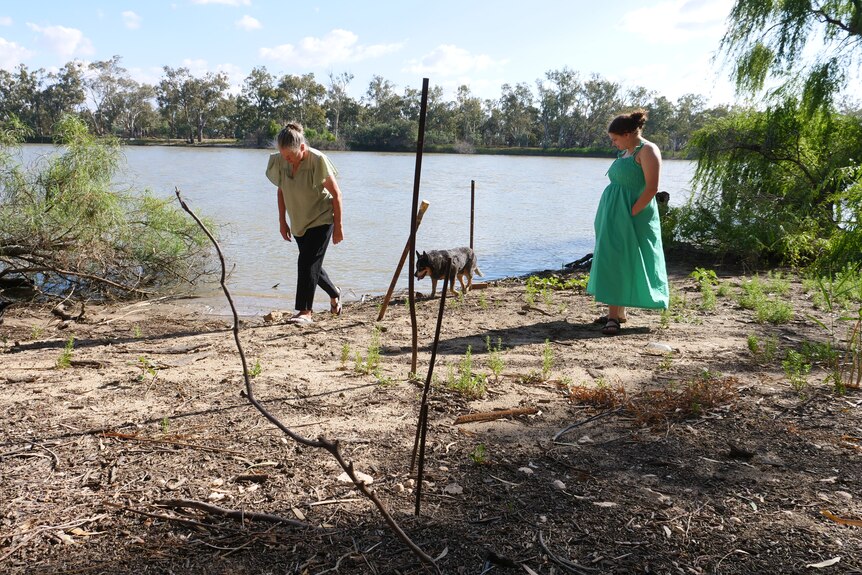 Dos mujeres paradas cerca del río, con palos en primer plano mostrando los niveles del agua.