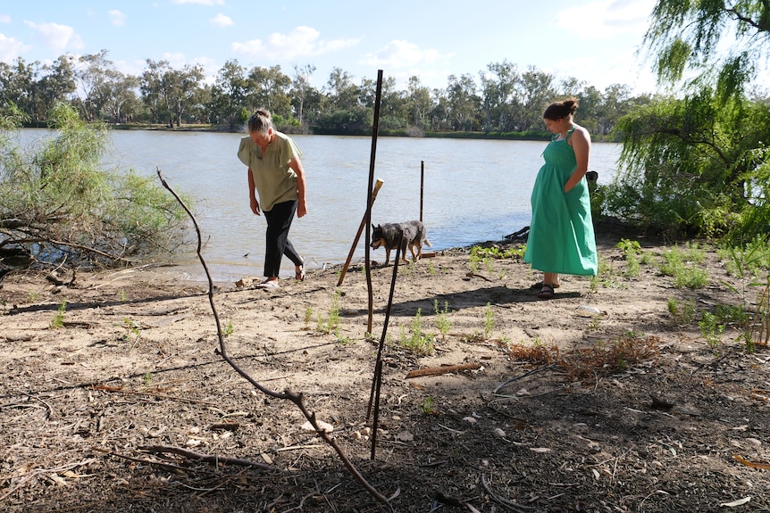 Two women standing near the river, with sticks in the foreground showing water levels.