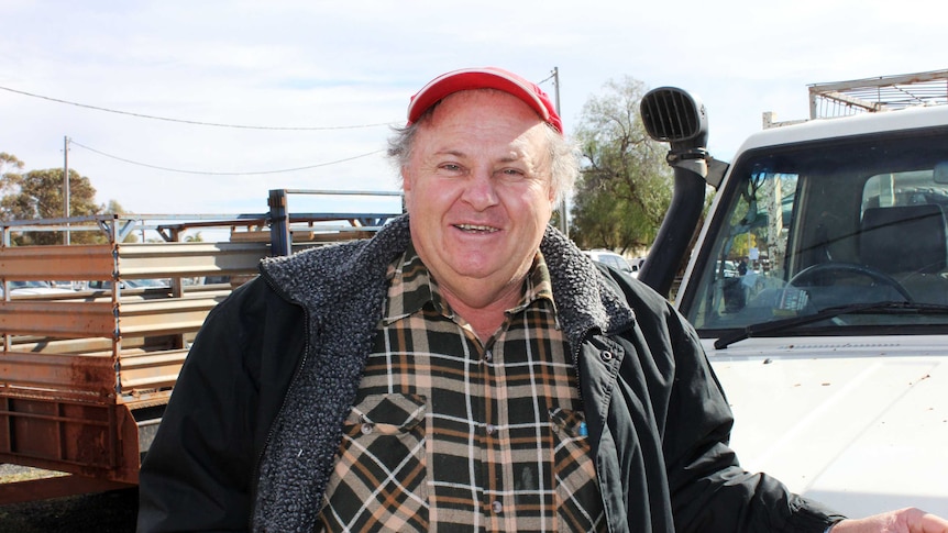 An older man in a red cap and warm clothes stands in front of a ute.
