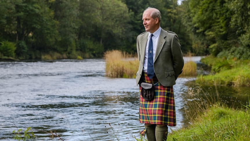 John Buchanan stands by a river wearing a tartan kilt, brown jacket and blue tie.