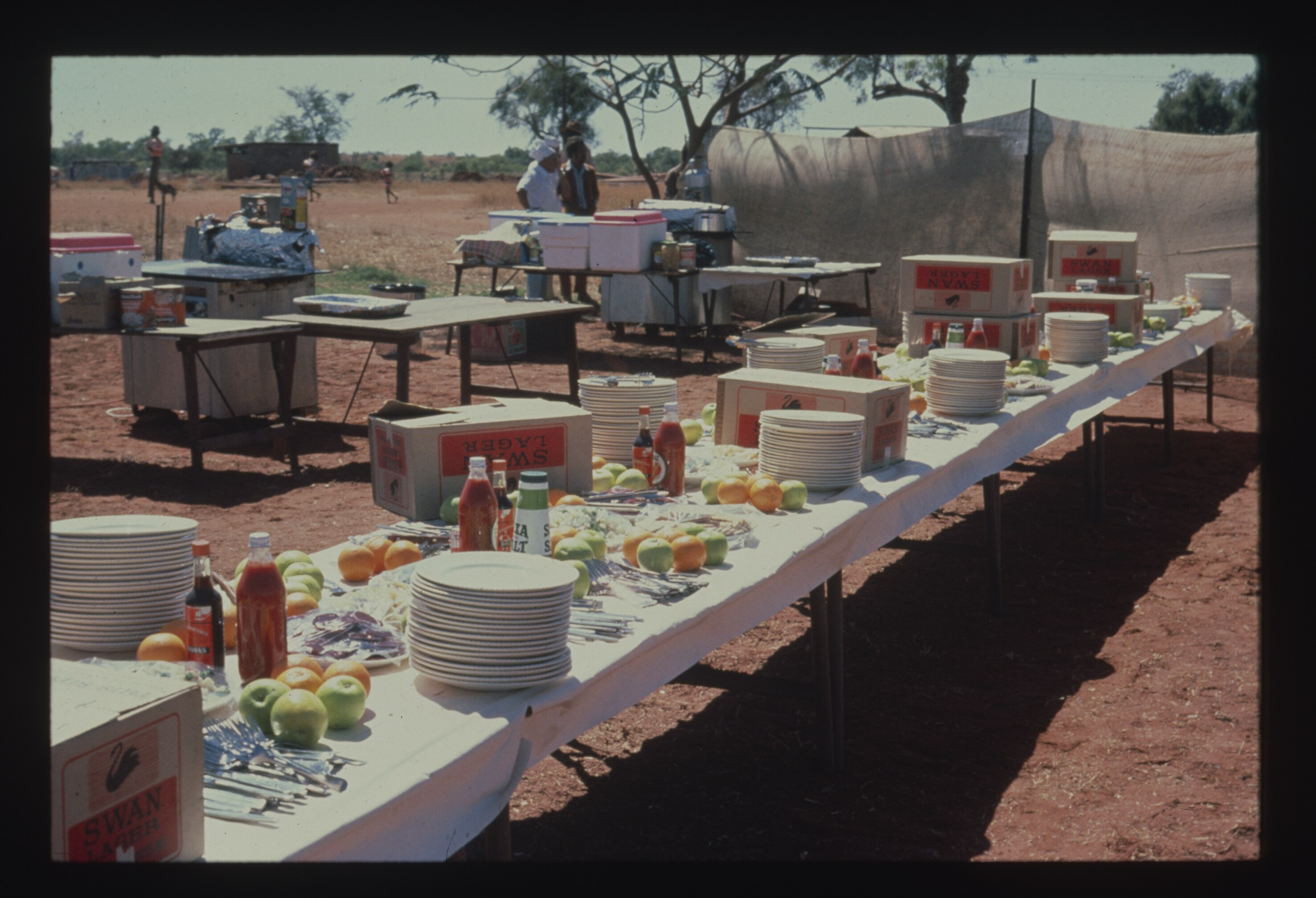Dated photo, long row of fold-out tables, with cloth, topped with plates and spread of food on dirt ground.