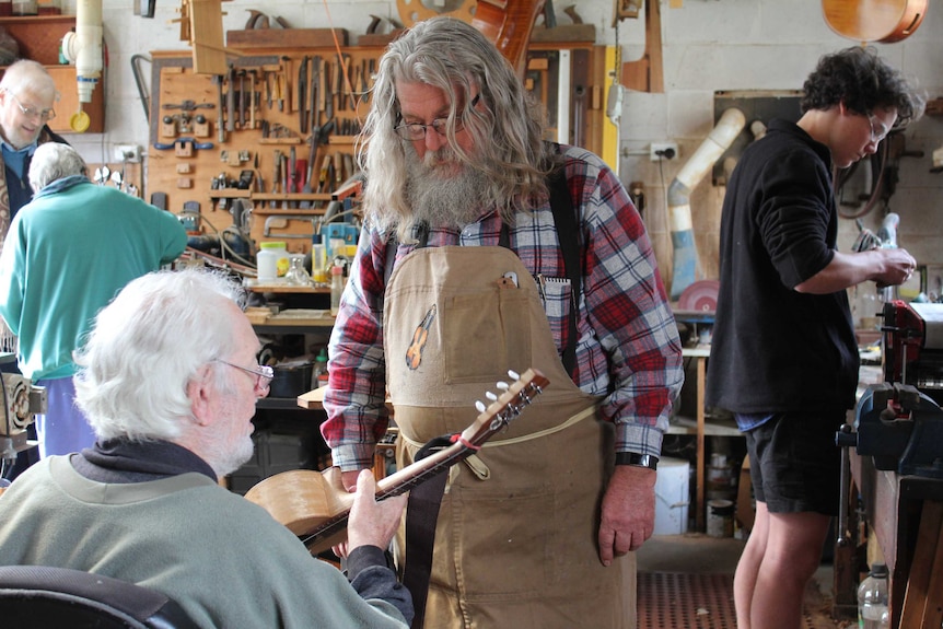 A man in an apron receives advice from an olded seated man holding a guitar in a busy workshop where other men are at work