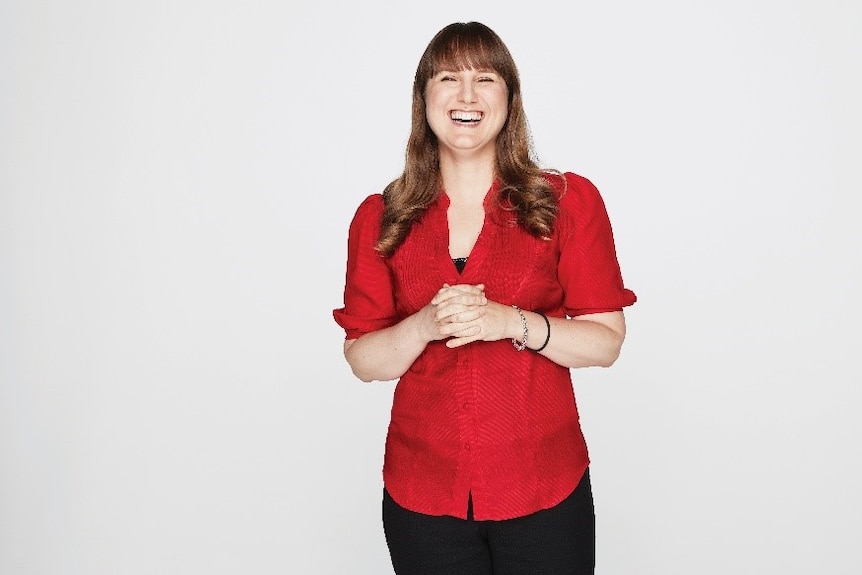 A woman stands against a white background, she is laughing and appears happy. 