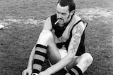 A footballer with a cigarette hanging in his mouth fiddles with his boots on the MCG.