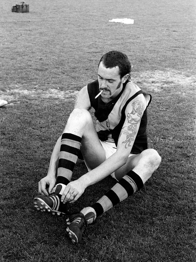 A footballer with a cigarette hanging in his mouth fiddles with his boots on the MCG.