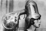 A vintage black and white photo of a large metal contraption drying a woman's hair.