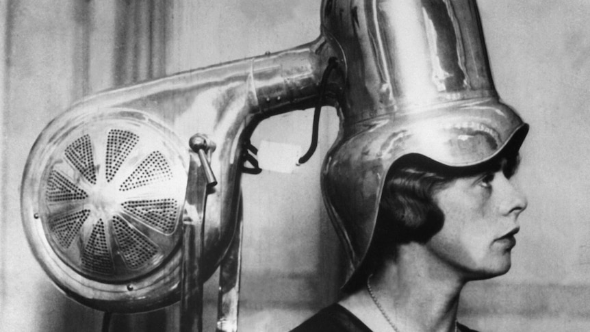 A vintage black and white photo of a large metal contraption drying a woman's hair.