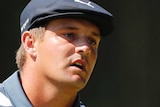 Bryson DeChambeau clenches his fist and looks forwards with his mouth partly open wearing a blue hat and striped polo shirt