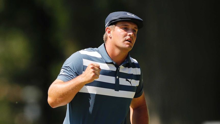 Bryson DeChambeau clenches his fist and looks forwards with his mouth partly open wearing a blue hat and striped polo shirt