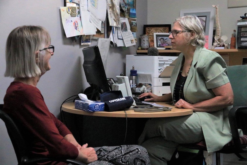 A female patient sits opposite a doctor at her desk in a GP clinic as the doctor looks at her computer.