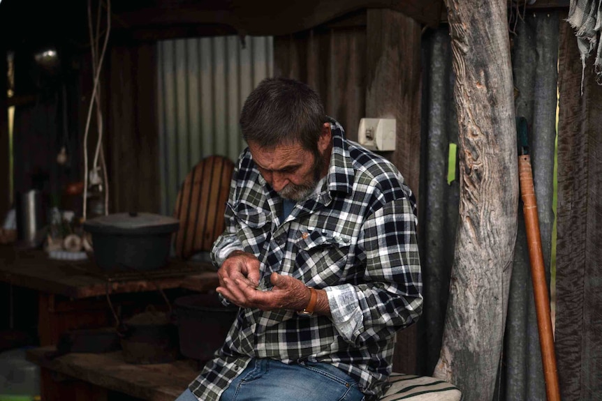 a man in a checked shirt looks at his hands in a shed.