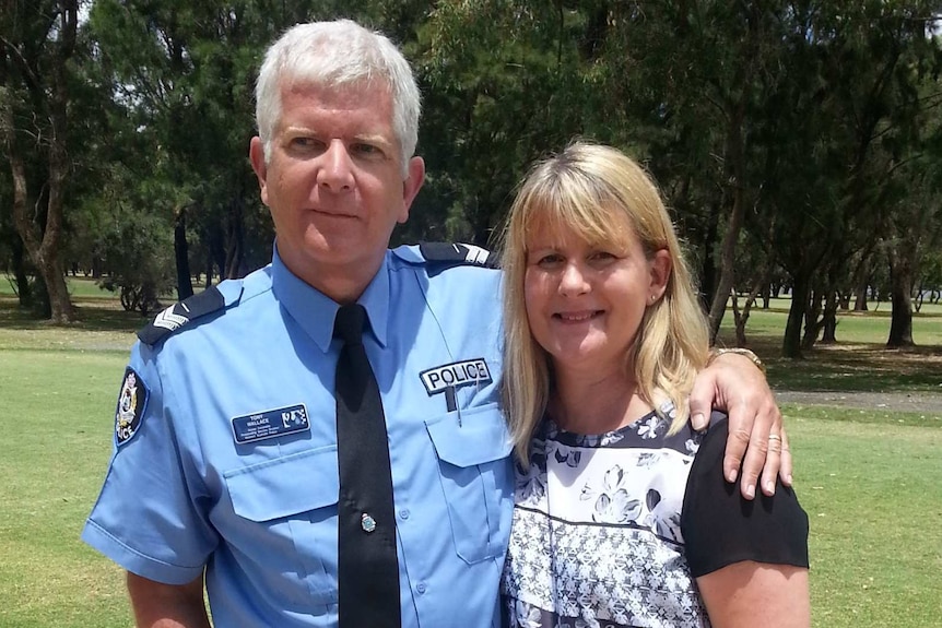 A man in police uniform stands with his arm around his wife in an outdoor location.