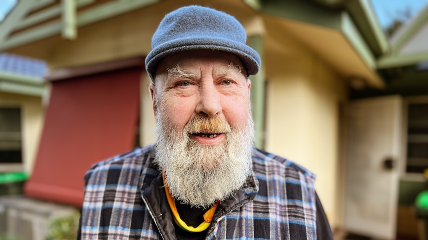 A man with a beard wearing a gray hat and a checked vest. His home is behind him.