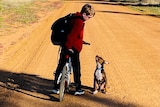 A boy with a red jumper stands on a bike and looks down at a little dog looking up at him.