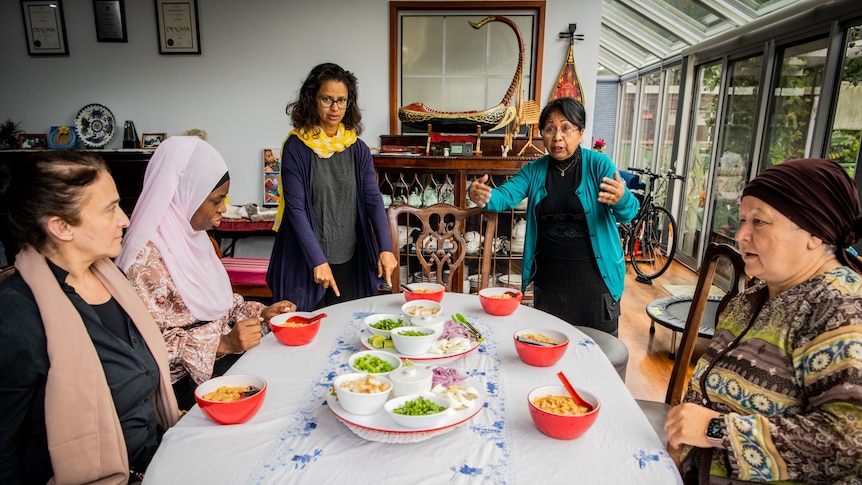 Five women around a dining table with Laksa soup in bright red bowls, Thit gestures as she talks to the group.