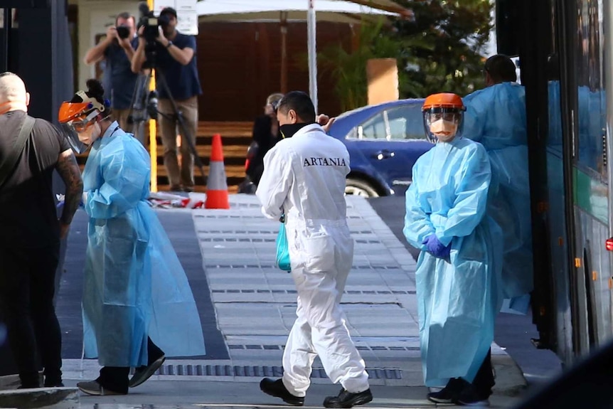 People in light blue and white protective clothing walk from a bus into a building.