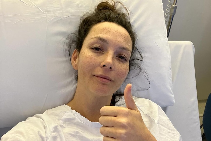 Singer Ricki Lee in a hospital bed and gown with her thumbs up