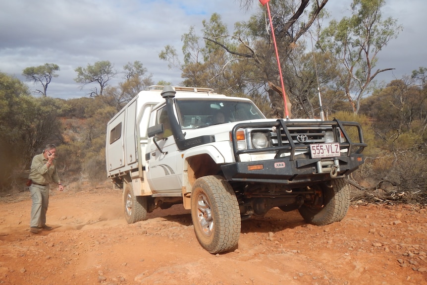 A man stand on the left with a small radio and a white car drives along a red dirt track. 