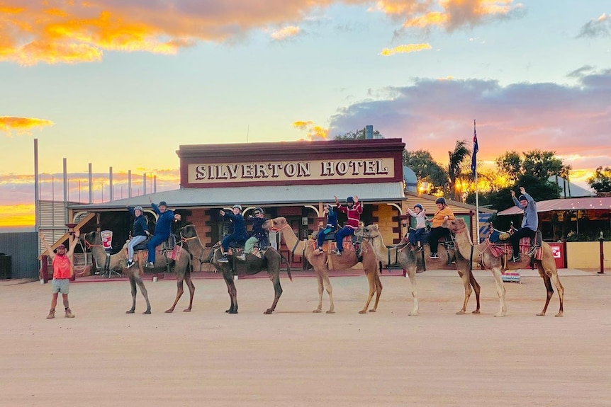 A man in a red shirt, leads a group of camels with people on them as the sun sets and the Silverton Hotel