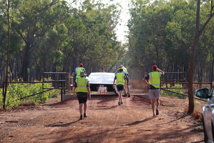 People wearing high vis walking away from the camera with a solar car along a dirt road in the bush.