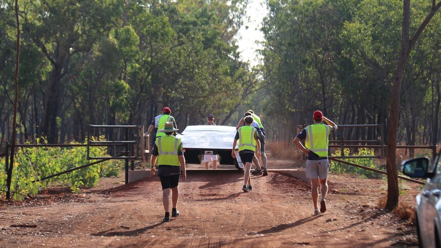 People wearing high vis walking away from the camera with a solar car along a dirt road in the bush.