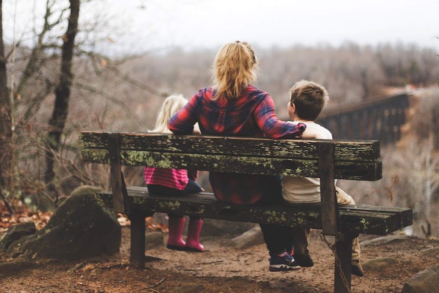 A woman sits on a park bench with her daughter and son.