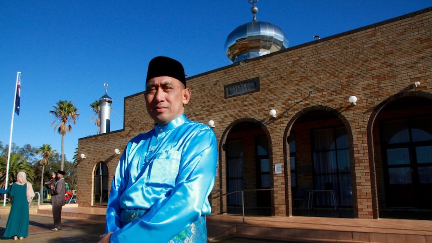 A muslim Imam in traditional dress standing outside mosque with Australian flag in the frame