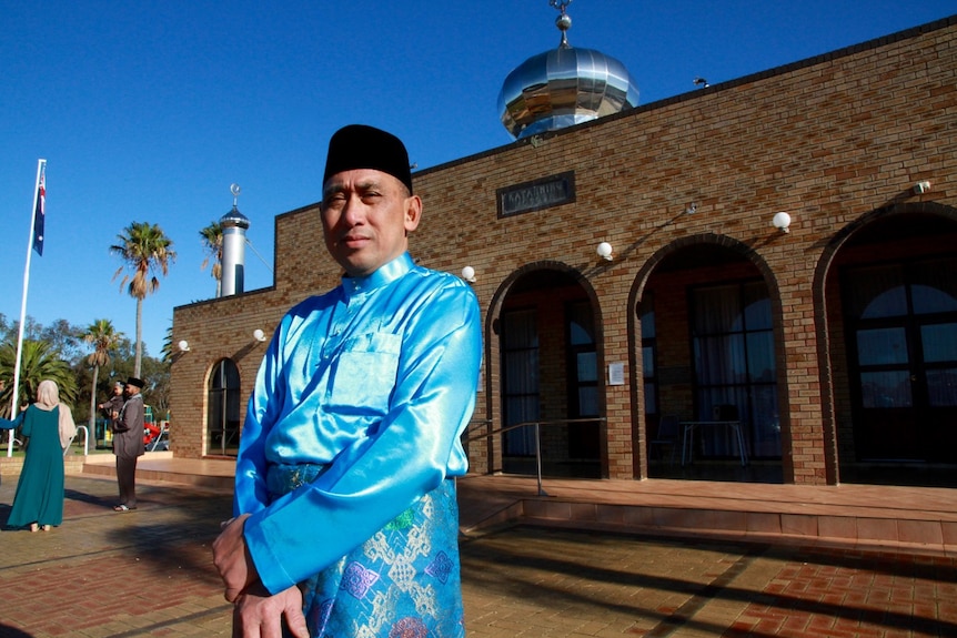 A muslim Imam in traditional dress standing outside mosque with Australian flag in the frame
