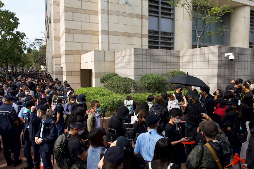 A large crowd of Chinese people wearing black and face masks stand around a tan coloured building.