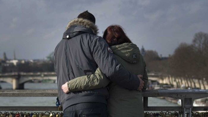 A couple with their backs to the camera embrace as they look at the view from a bridge.
