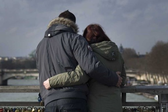 A couple with their backs to the camera embrace as they look at the view from a bridge.