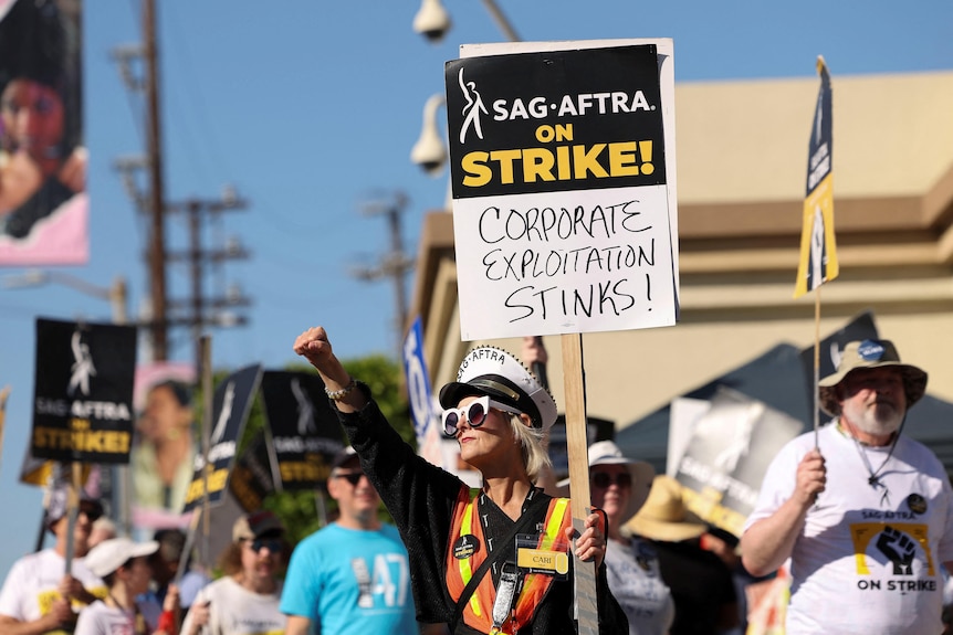 Striking SAG AFTRA member holds up sign saying corporate exploitation stinks 