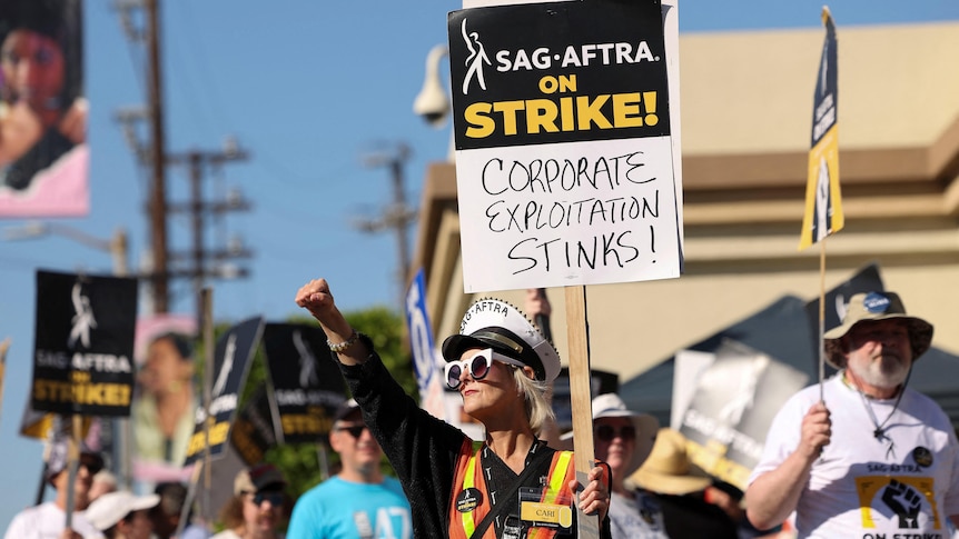 Striking SAG AFTRA member holds up sign saying corporate exploitation stinks 