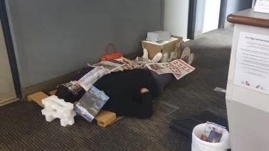 Man lies on floor with newspapers spread over him as blanket