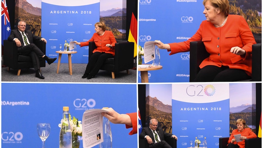 Angela Merkel sitting at a table with Scott Morrison. In Ms Merkel's hand is a piece of a paper with a photo of Mr Morrison.