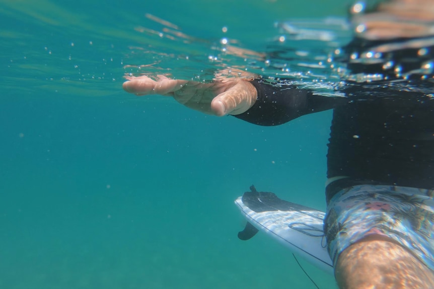 An underwater image of a hand half-submerged. Part of surfer sitting on board in background